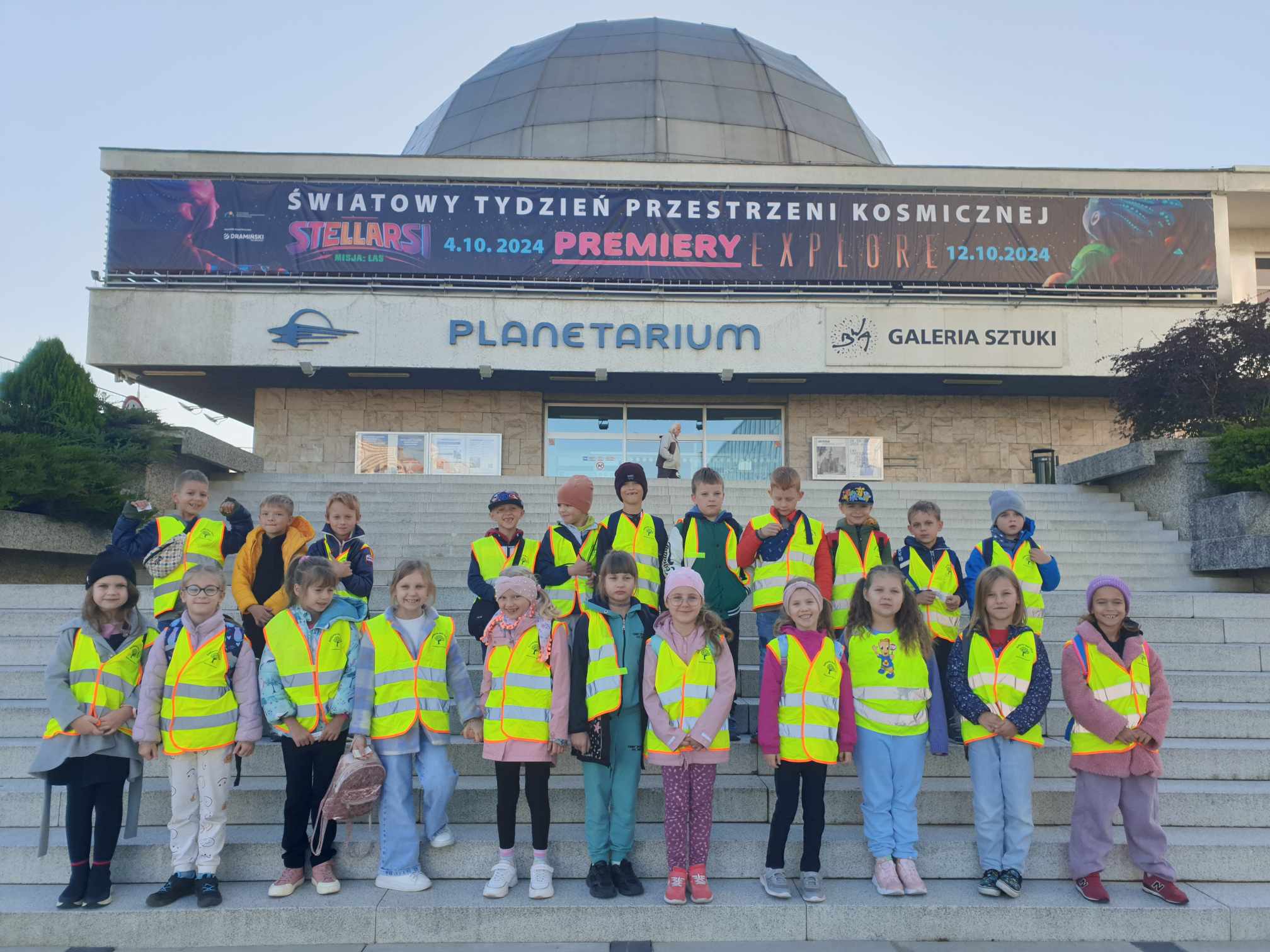 Na zdjęciu widzimy grupę dzieci stojących na schodach przed planetarium. Dzieci ubrane są w żółte odblaskowe kamizelki, co sugeruje, że są na wycieczce szkolnej. W tle znajduje się budynek planetarium z napisem promującym wydarzenie związane z kosmosem, odbywające się w październiku 2024 roku. Baner nad wejściem zapowiada światowy tydzień przestrzeni kosmicznej oraz premiery filmowe. Dzieci wydają się być w dobrych nastrojach, a pogoda jest słoneczna, co sprzyja zwiedzaniu.