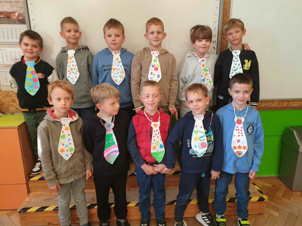 Na zdjęciu widać grupę jedenastu chłopców, którzy stoją w dwóch rzędach na tle tablicy. Wszyscy mają na sobie własnoręcznie wykonane, ozdobione papierowe krawaty na sznurkach, które są kolorowe i różnorodnie ozdobione, na przykład w kwiatki, gwiazdki i inne wzory. Chłopcy mają na sobie codzienne ubrania, takie jak bluzy z kapturem i koszulki, a ich twarze wyrażają zadowolenie i uśmiech. Tło stanowi szkolna klasa z białą tablicą i widocznym fragmentem kalendarza oraz zieloną ścianą. Chłopcy dostali krawaty od dziewczynek z okazji Dnia Chłopaka.