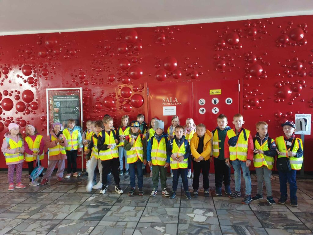 Na zdjęciu widzimy grupę dzieci ubranych w odblaskowe kamizelki, stojących na tle czerwonej ściany. Dzieci wyglądają na gotowe do wycieczki lub zwiedzania, są ustawione w szeregu, patrząc w kierunku aparatu. W tle znajduje się ściana pokryta wypukłymi, okrągłymi elementami, a na drzwiach widnieje napis „Sala Projekcyjna”. Na ścianie można także dostrzec kilka znaków, które najprawdopodobniej informują o zasadach panujących w tym miejscu. Całość sprawia wrażenie, że zdjęcie zostało zrobione w planetarium.