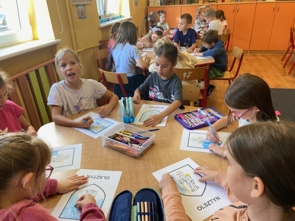 Na zdjęciu widać dzieci siedzące przy stolikach. Dzieci kolorują herb Olsztyna.