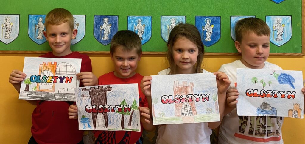 Na zdjęciu widać czworo dzieci trzymające prace plastyczne z wizerunkiem zabytków olsztyńskich. Na środku napis Olsztyn. W tle zielona tablica z herbami Olsztyna namalowanymi przez dzieci.