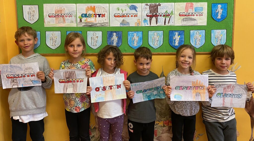 Na zdjęciu widać sześcioro dzieci trzymające prace plastyczne z wizerunkiem zabytków olsztyńskich. Na środku napis Olsztyn. W tle zielona tablica z herbami Olsztyna oraz zabytkami olsztyńskimi namalowanymi przez dzieci.
