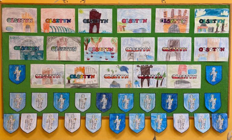 Zielona tablica z wystawą prac namalowanymi przez dzieci: herbami Olsztyna oraz zabytkami olsztyńskimi.