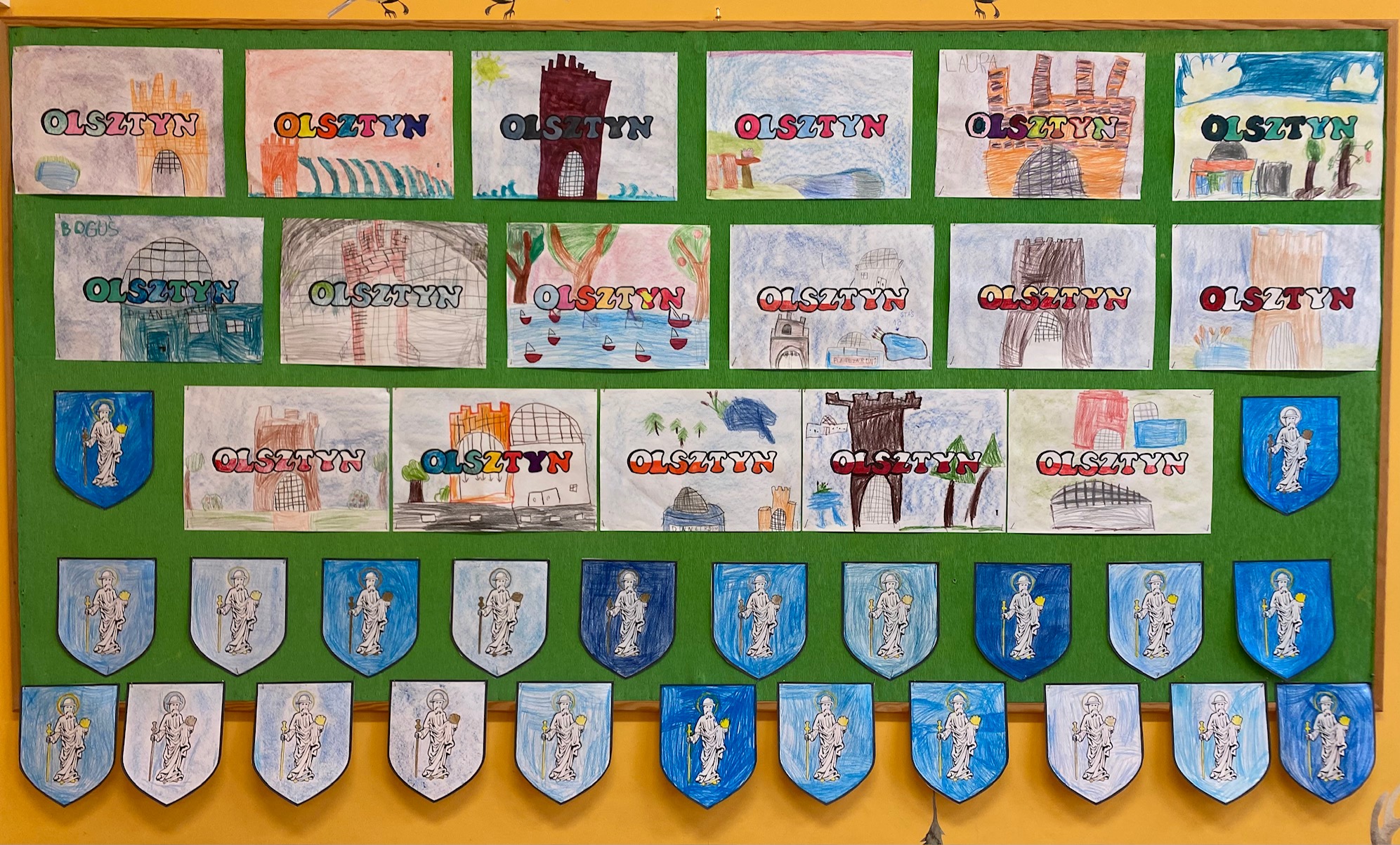 Zielona tablica z wystawą prac namalowanymi przez dzieci: herbami Olsztyna oraz zabytkami olsztyńskimi.