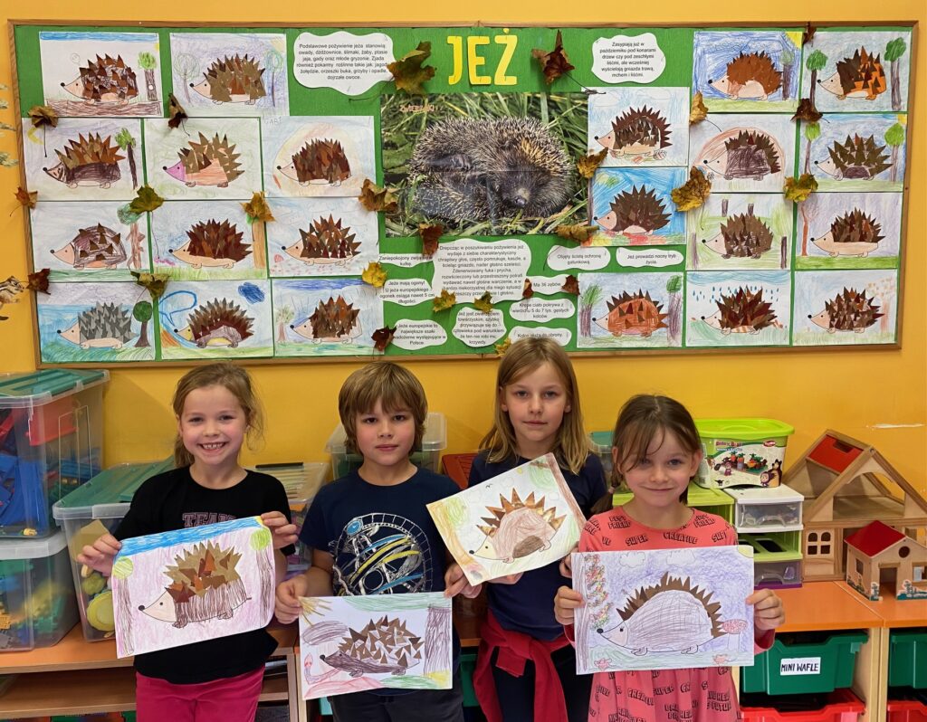 Na zdjęciu widać czworo dzieci trzymające swoje prace plastyczne przedstawiające jeża z kolcami naklejonymi z papieru kolorowego. W tle widać tablicę z informacjami o jeżu, zdjęciem jeża oraz pracami plastycznymi dzieci, przedstawiającymi jeża.
