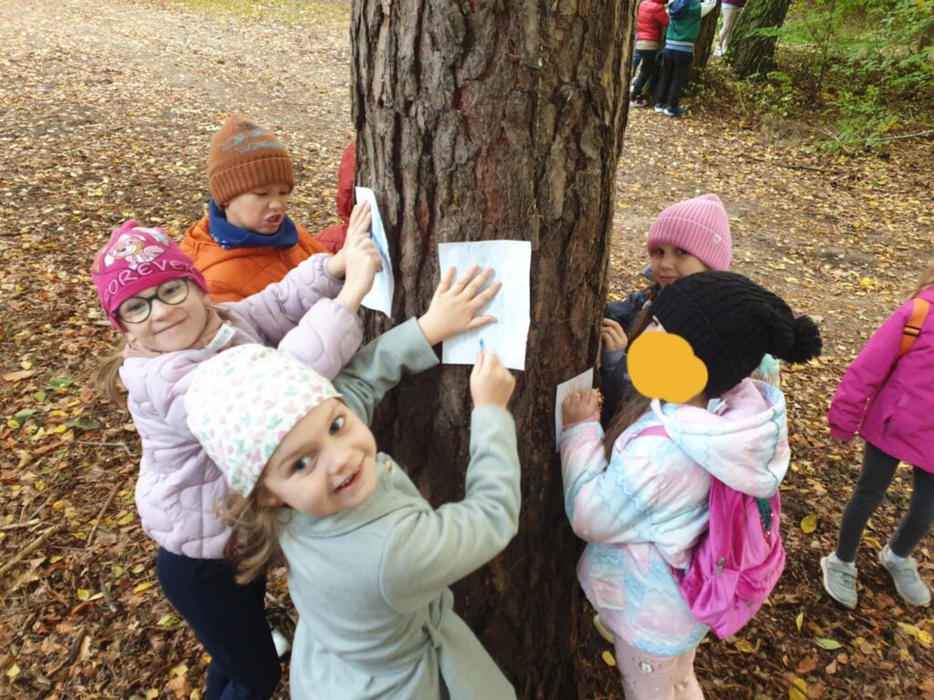 Na zdjęciu widzimy grupkę dzieci bawiących się przy drzewie. Dzieci trzymają kartki papieru przy pniu drzewa, prawdopodobnie robiąc odciski kory. Są ubrane w ciepłe kurtki i czapki, co sugeruje chłodniejszą porę roku. Dzieci wyglądają na zadowolone i skupione na swojej zabawie. W tle widać inne dzieci, które również biorą udział w aktywnościach na świeżym powietrzu.