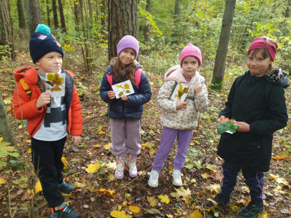 Na zdjęciu widać czwórkę dzieci stojących w lesie, każde z nich trzyma w rękach kartkę z przykładem jesiennych liści. Ubrane są w ciepłe ubrania – czapki, kurtki, a na ziemi widać kolorowe, opadłe liście. Dzieci wyglądają na zadowolone, uśmiechają się i wydaje się, że biorą udział w jakiejś jesiennej zabawie lub lekcji przyrody. W tle znajduje się las, pełen zielonych i żółtych liści, co świadczy o jesiennej porze roku. Jest to wycieczka edukacyjna, podczas której dzieci uczą się o przyrodzie.
