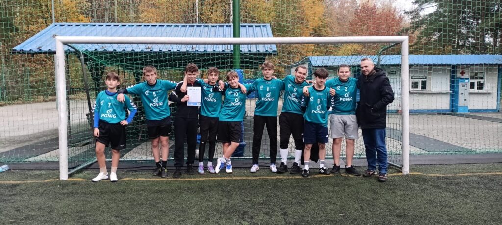 Na zdjęciu znajduje się grupa młodych sportowców oraz ich trener, ustawieni przed bramką na boisku piłkarskim. Chłopcy mają na sobie zielone koszulki z logo „PGNiG” i różne numery na koszulkach, co wskazuje na drużynę sportową. Wszyscy są ubrani na sportowo, gotowi do gry lub po zakończonym meczu. Jeden z chłopców trzyma dyplom, prawdopodobnie za osiągnięcie drużyny. W tle widać siatkę bramki oraz budynki z niebieskim dachem, a za nimi jesienne drzewa w odcieniach żółci i pomarańczu, co sugeruje, że zdjęcie zostało zrobione w sezonie jesiennym.