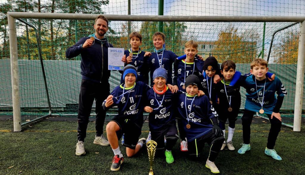 Na zdjęciu widzimy młodą drużynę sportową, wraz z trenerem, ustawioną przed bramką na boisku piłkarskim. Wszyscy zawodnicy mają na sobie granatowe stroje z logo „PGNiG” oraz numery na koszulkach. Każdy z chłopców nosi medal, co sugeruje, że drużyna zdobyła miejsce na podium w niedawnym turnieju. Trener, stojący po lewej stronie, trzyma dyplom, a przed grupą widoczny jest puchar. W tle widać siatkę bramki oraz drzewa, których liście przybrały jesienne kolory, co sugeruje, że zdjęcie wykonano jesienią.