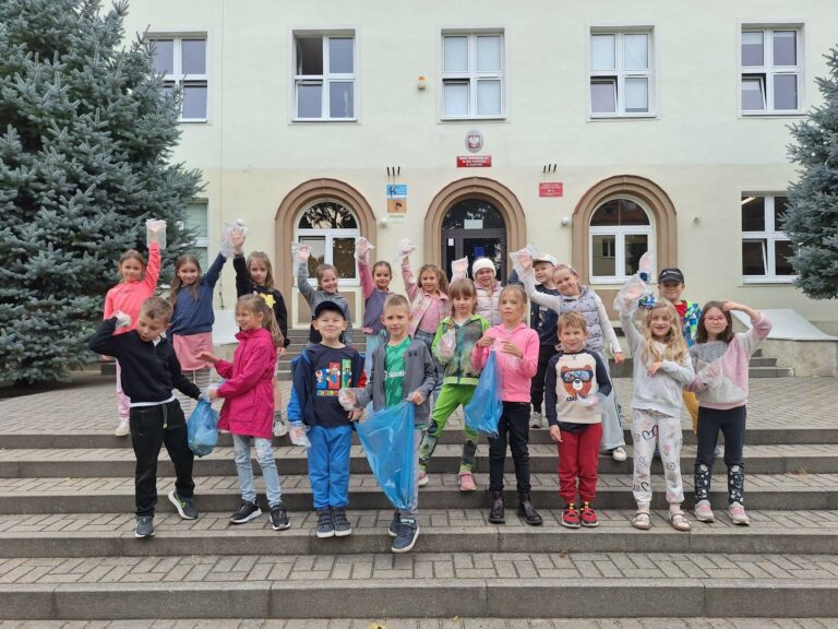 Na zdjęciu znajduje się grupa około 20 dzieci stojących na schodach przed budynkiem szkoły. Większość dzieci jest uśmiechnięta i pozuje z plastikowymi rękawiczkami, trzymając w dłoniach worki na śmieci, co sugeruje udział w akcji sprzątania. W tle widać dwa wysokie świerki oraz front budynku szkoły z dużymi oknami i trzema łukowatymi drzwiami. Dzieci są ubrane na sportowo w kolorowe bluzy, kurtki i spodnie, a ich wiek to prawdopodobnie 6-10 lat. Atmosfera jest radosna, a dzieci wyglądają na zaangażowane w wydarzenie.