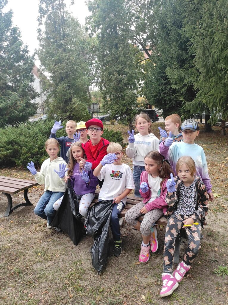 Cztery dziewczynki i jeden chłopiec siedzą na ławce. Dwoje dzieci trzyma worki na śmieci. Za nimi stoi 5 chłopców i 1 dziewczynka. Trzech chłopców ma czapki na głowie.