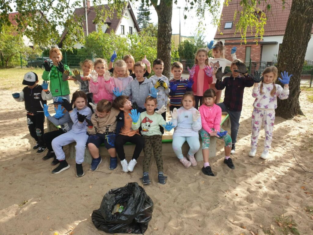 Na zdjęciu znajduje się grupa dzieci, które uczestniczą w akcji sprzątania. Wszystkie dzieci mają na dłoniach kolorowe rękawiczki ochronne – niebieskie, czarne, białe, i różowe. Siedzą i stoją w grupie na piasku, na placu zabaw, a przed nimi znajduje się worek na śmieci z odpadami. W tle widać domy z ogrodami, co sugeruje, że akcja odbywa się na terenie osiedla. Dzieci wydają się być wesołe i zadowolone ze wspólnej inicjatywy.
