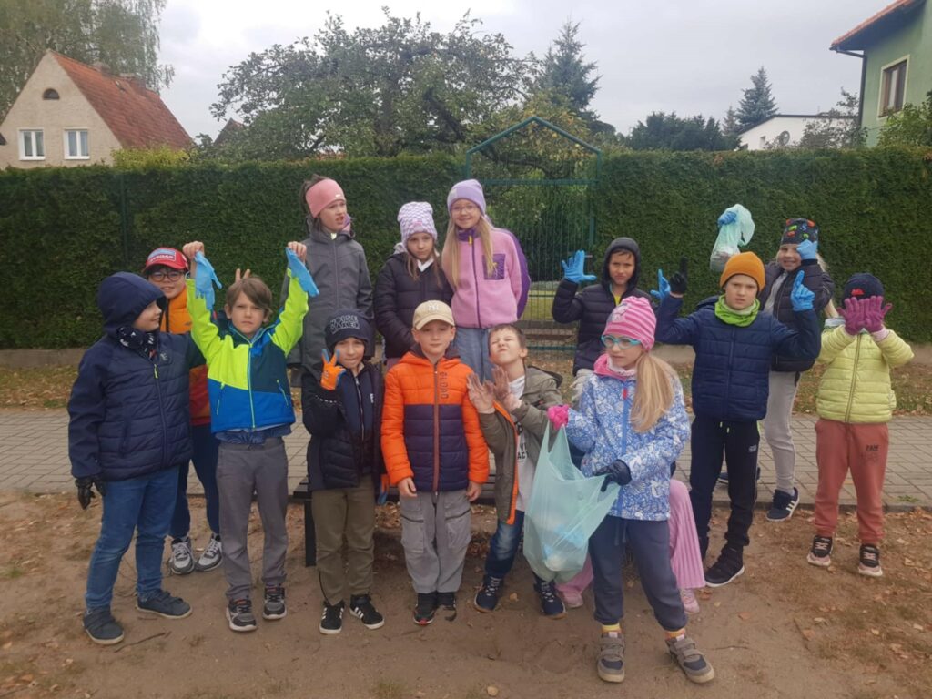Na podwórku stoi kilkunastoosobowa grupa dzieci w kurtkach. Część dzieci podniosła do góry ręce w rekawiczkach jednorazowych. Z przodu stpoi dziewczunka i trzyma niebieski worek. Dzieci uczestniczyły w akcji sprzątania terenu.