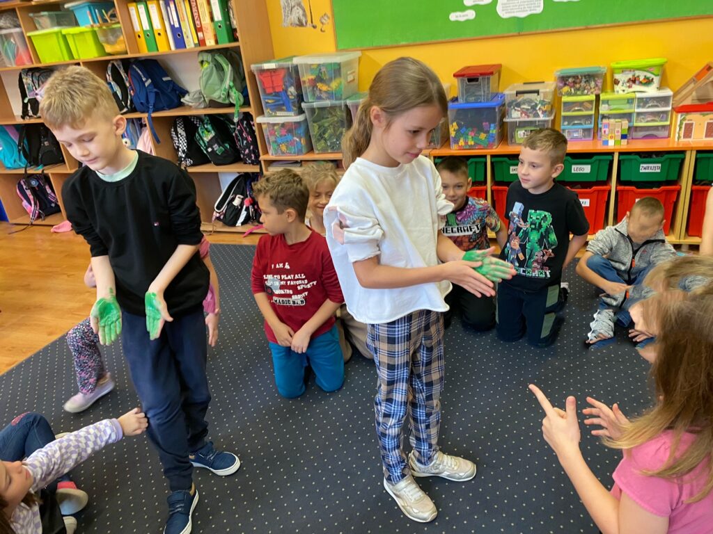 Dzieci siedzą w kole. Chłopiec i dziewczynka stojący w środku koła pokazują jak dokładnie rozprowadzić mydło (farbę)na dłoniach.