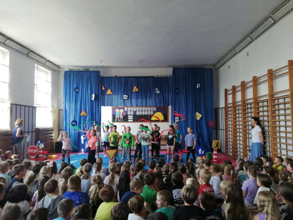 Na zdjęciu widać salę gimnastyczną. Główna ściana udekorowana jest niebieskimi zasłonami, na nich przyczepione sa różne znaki drogowe. W głębi widać tablicę z napisem i sylwety dzieci i auta. Przed dekoracjami stoi grupa dzieci, która występuje przed publicznością. Dzieci trzymaja szarfy zielone i czerwone, śpiewaja piosenkę. Na podłodze widać siedzące dzieci, jest ich bardzo dużo- to widownia. Po bokach sali stoją 2 nauczycielki.