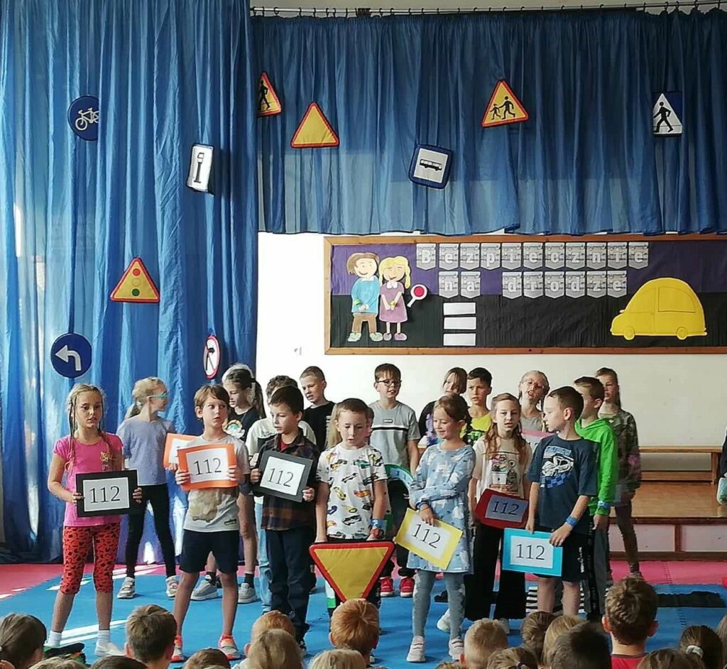 Na tle dekoracji ( niebieskie zasłony z przypiętymi znakami drogowymi oraz tablica z napisem i sylwetkami dzieci i auta) stoi grupa dzieci. W rękach trzymają tabliczki z numerem 112 i śpiewają piosenkę.