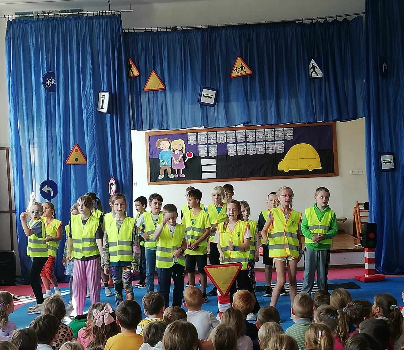 Na tle dekoracji ( niebieskie zasłony z przypiętymi znakami drogowymi oraz tablica z napisem i sylwetkami dzieci i auta) stoi grupa dzieci. Są ubrane w żółte, odblaskowe kamizelki. Niektóre dzieci mają podniesione ręce. Dzieci śpiewają i tańczą. Przed nimi widać fragment publiczności.