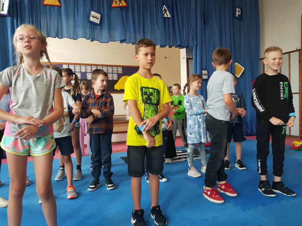 Cały kadr zajmuje grupka dzieci (ok.10 osób). Stoją na niebieskiej podłodze i na tle niebieskiego materiału. Na ręce mają założone odblaski i śpiewają piosenkę.