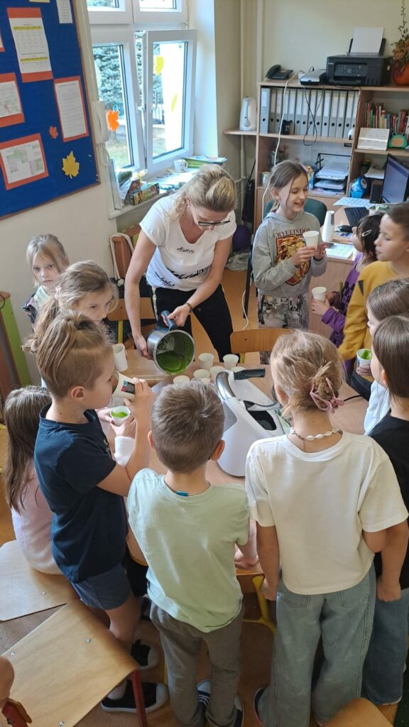 11 dzieci stoi wokół stolika. 8 dziewczynek i 3 chłopców. Nauczyciel wlewa z dzbanka metalowego koktajl zrobiony w robocie kuchennym do kubeczków pierowych 200 ml. Koktajl został wykonany