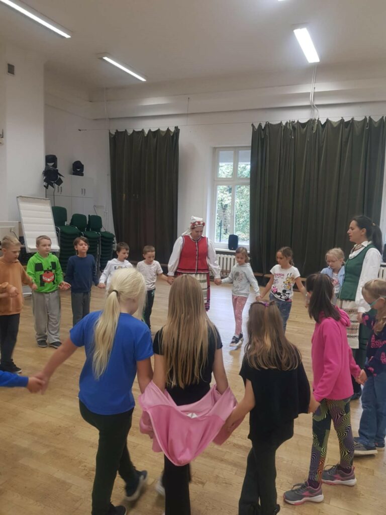 W przestronnej sali, grupa dzieci stoi w kole, trzymając się za ręce. Wśród nich sa dwie dorosłe kobiety w warmińskich strojach. Uczą dzieci kroków tańca.