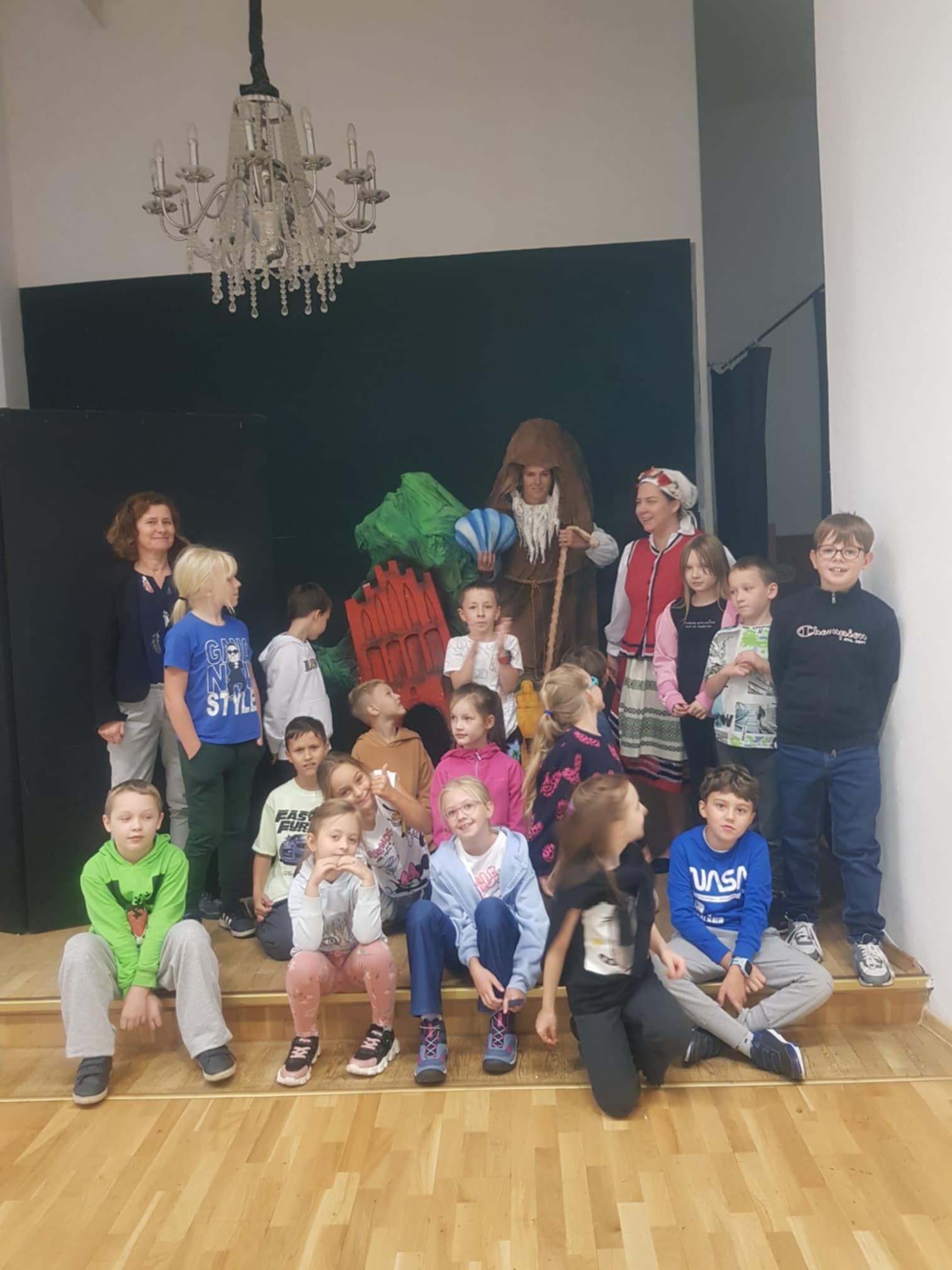 W sali, na podwyższonym stopniu siedzi kilkoro dzieci. Za nimi stoją kolejne dzieci oraz trzy dorosłe kobiety. Jedna z nich ubrana jest w strój warmiński. Grupa ustawiła się w ten sposób do zdjęcia.