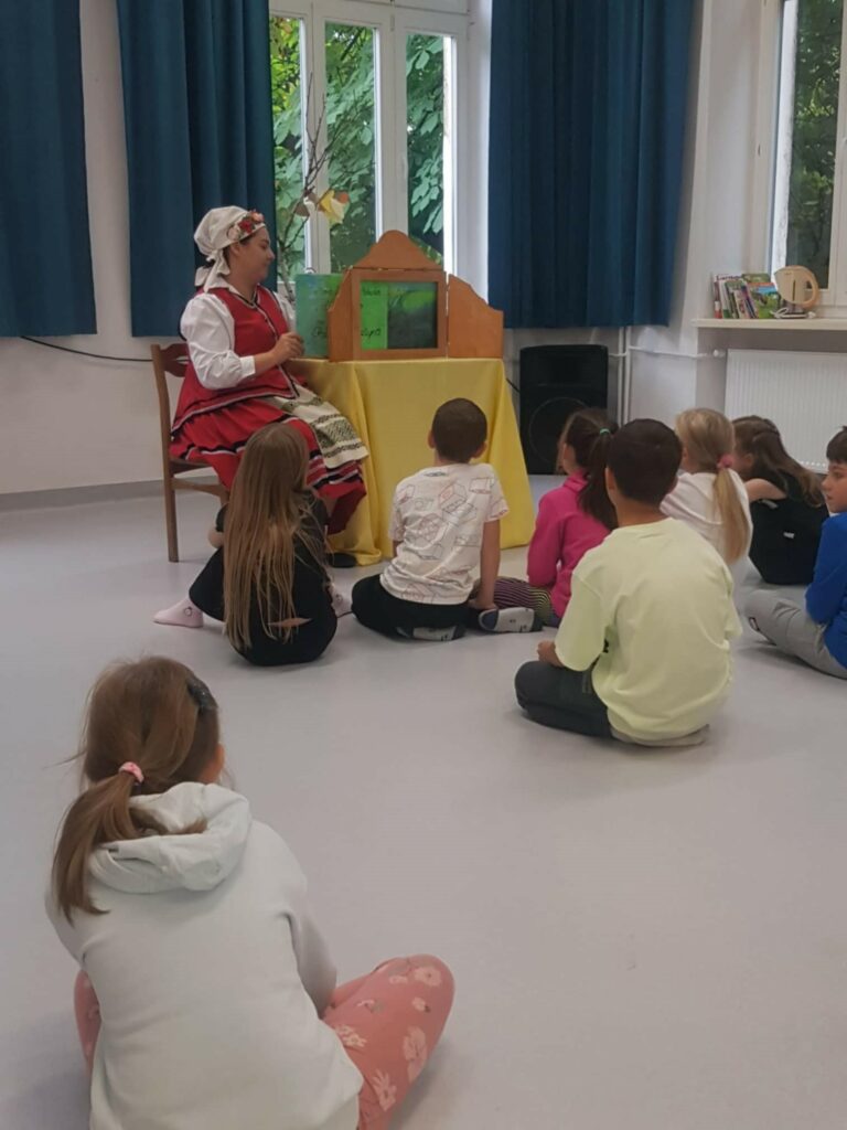 W sali, na tle okna, siedzi pani w stroju warmińskim. Obok, na stoliku stoi teatr kamishibai. Twarzą do teatrzyku siedzą dzieci. Na zdjęciu widać plecy dzieci.