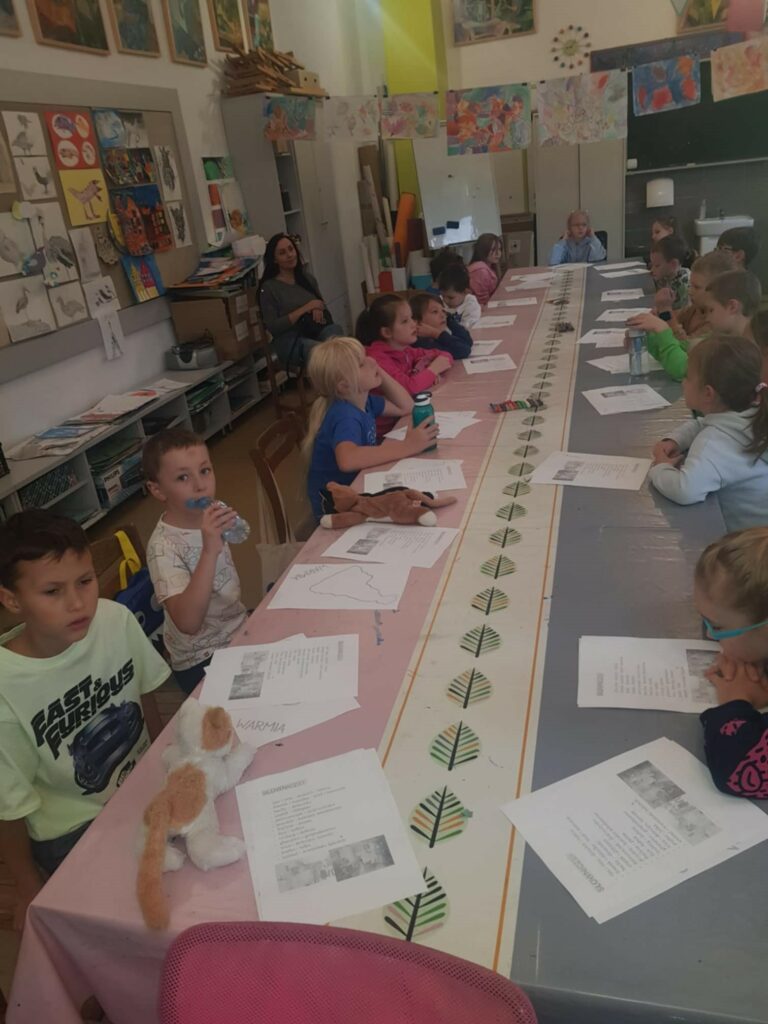 Na zdjęciu dzieci siedzą przy stole, trzymając kartki papieru podczas zajęć, a nauczycielka stoi z przodu, prowadząc lekcję. W tle widać jasną salę z oknami i kolorowymi pracami plastycznymi zawieszonymi na ścianach.