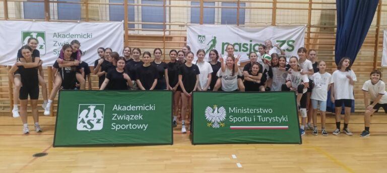 Zdjęcie przedstawia dużą grupę dzieci i młodzieży uczestniczących w wydarzeniu sportowym w sali gimnastycznej. Uczestnicy są różnorodnie ubrani – część z nich ma czarne stroje sportowe, a inni białe koszulki. Grupa pozuje w radosnej atmosferze, niektórzy stoją, inni trzymają się na barana lub siedzą na podłodze. W tle widoczne są banery z napisami: “Akademicki Związek Sportowy” oraz “Ministerstwo Sportu i Turystyki”, co sugeruje, że wydarzenie jest organizowane w ramach programu promującego sport i aktywność fizyczną. Atmosfera na zdjęciu jest luźna i pełna energii, co wskazuje na integrację i dobrą zabawę podczas zajęć.