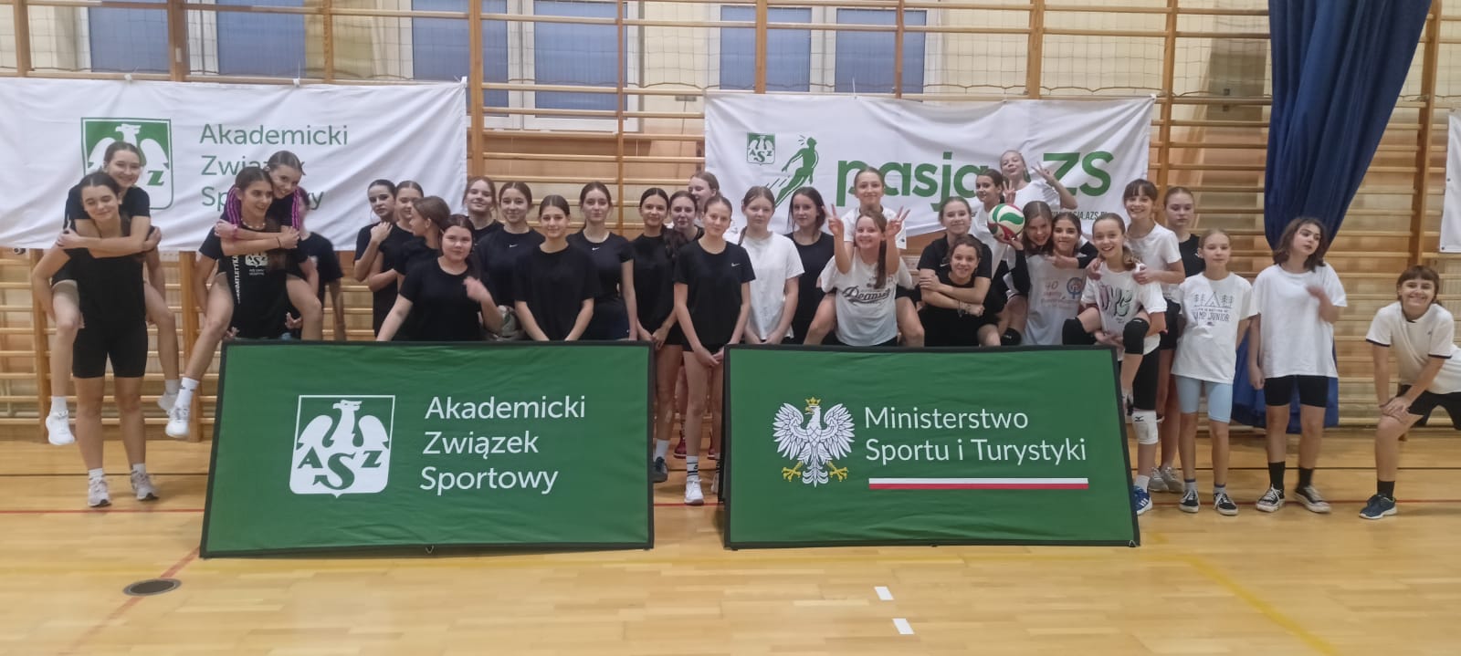 Zdjęcie przedstawia dużą grupę dzieci i młodzieży uczestniczących w wydarzeniu sportowym w sali gimnastycznej. Uczestnicy są różnorodnie ubrani – część z nich ma czarne stroje sportowe, a inni białe koszulki. Grupa pozuje w radosnej atmosferze, niektórzy stoją, inni trzymają się na barana lub siedzą na podłodze. W tle widoczne są banery z napisami: “Akademicki Związek Sportowy” oraz “Ministerstwo Sportu i Turystyki”, co sugeruje, że wydarzenie jest organizowane w ramach programu promującego sport i aktywność fizyczną. Atmosfera na zdjęciu jest luźna i pełna energii, co wskazuje na integrację i dobrą zabawę podczas zajęć.