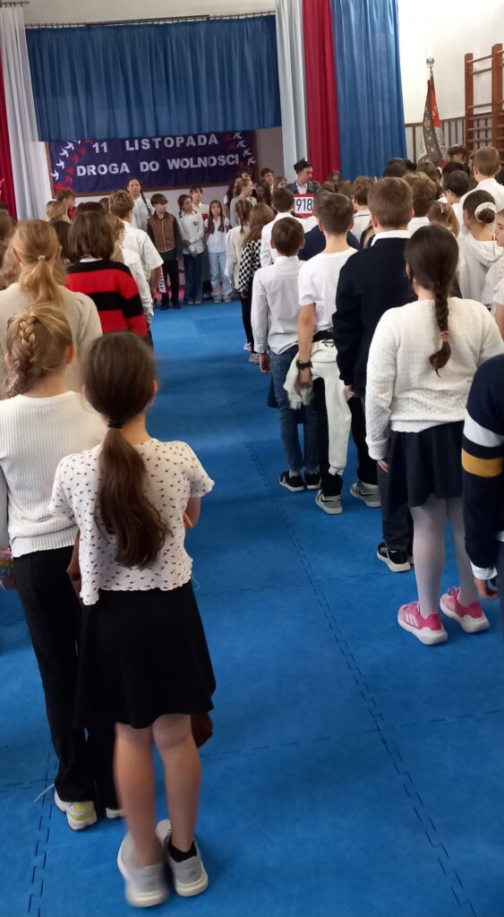 czniowie w pozycji baczność śpiewają “Mazurka Dąbrowskiego”, zwróceni przodem do sztandaru szkoły, a tyłem do fotografującego. W tle ściana z tablicą i napisem:"11 listopada - Droga do wolności" i zawieszonym materiałem w kolorze niebieskim oraz białe i czerwone szerokie szarfy.