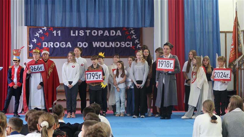Grupa uczniów stoi w szeregu. Kilka osób ma na sobie stroje księcia, biskupa, kadeta, Piłsudskiego. W rękach trzymają kartki z datami.W tle widać napis 11. listopada drogo do wolności. Wokół grupy widać niebieskie zasłony a na nich długie pasma tkaniny upięte w biało czerwoną flagę.
