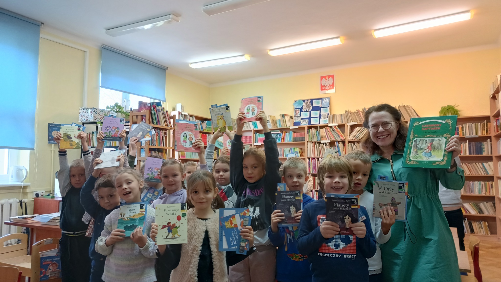 Zdjęcie przedstawia grupę dzieci chłopców i dziewczęta trzymających w dłoniach książki. Część dzieci trzyma książki nad głową część przed sobą. Obok grupy stoi uśmiechnięta nauczycielka również z książką w dłoni. W tle widać regały z książkami sugerujące że wydarzenia ma miejsce w bibliotece. Nad regałami widać godło Polski.