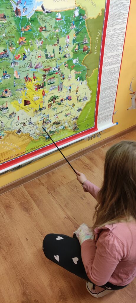zdjęcie przedstawia wiszącą na ścianie mapę Polski i siedzącą przed mapą dziewczynkę, która pokazuje wskaźnikiem Kraków na mapie.