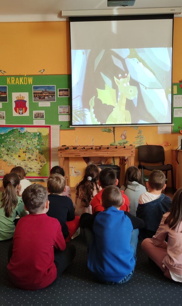 zdjęcie przedstawia siedzącą na granatowym dywanie grupę dzieci, które oglądają na ekranie ściennym z rzutnika postać Smoka Wawelskiego z legendy. Obok ekranu widać tablicę ścienną z zielonym tłem na której są umieszczone informacje o Krakowie. Pod tablicą wisi mapa Polski.