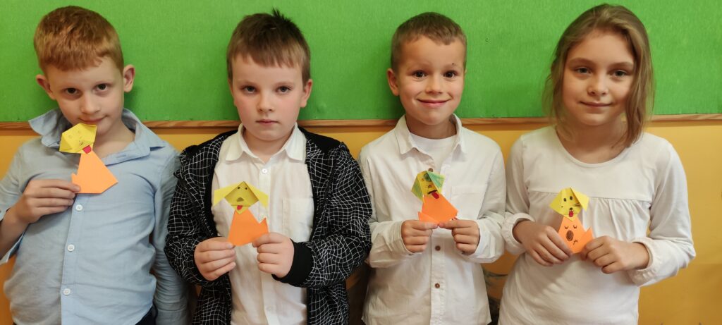 zdjęcie przedstawia grupę stojących dzieci pod zielona tablicą - 3 chłopców i 1 dziewczynkę. Dzieci trzymają w rękach wykonane z pomarańczowego i żółtego papieru podobizny piesków. Są one wykonane metodą origami.