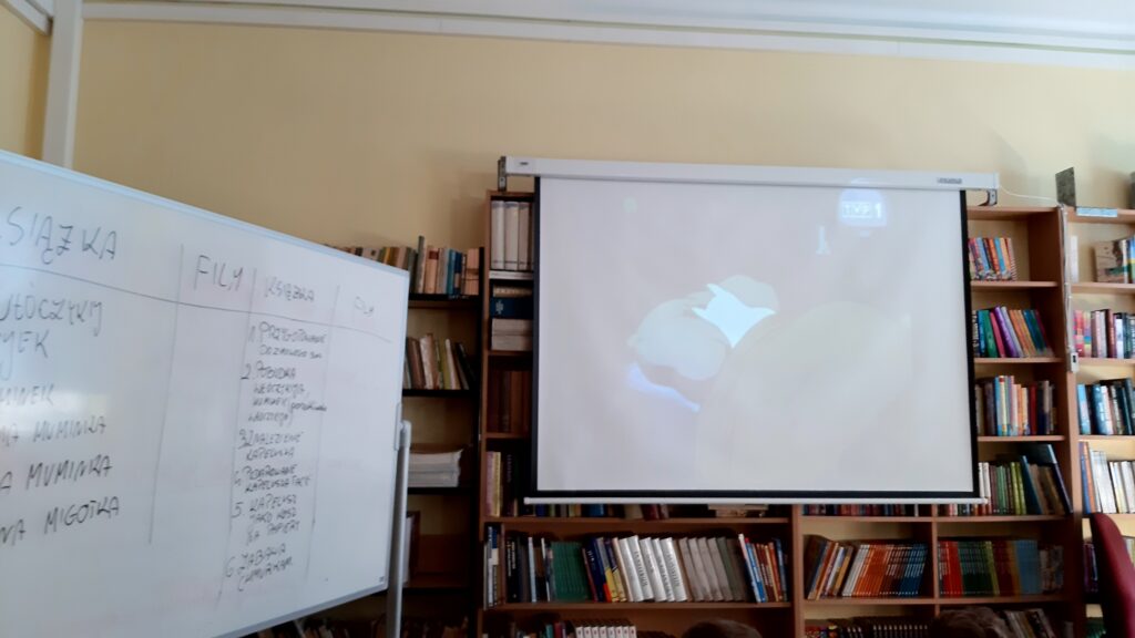 Zdjęcie przedstawia biały ekran rozciągnięty na tle półek z książkami. Na ekranie widac wyświetlaną animację. Z prawej strony zdjęcia widać stojąca do fotografa lekko bokiem białą zapisaną tablicę.