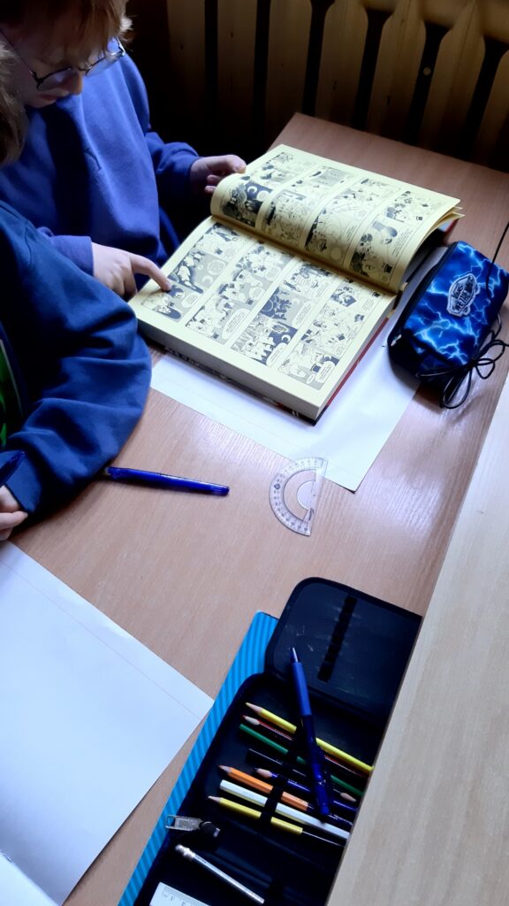 Zdjęcie przedstawia ławkę szkolną przy której siedzą uczniowie. Jeden ma przed sobą otwartą grubą książkę jest to komiks. Drugiego ucznia widać tylko łokieć położony na ławce, przed nim widać białą kartkę i otwarty piórnik.