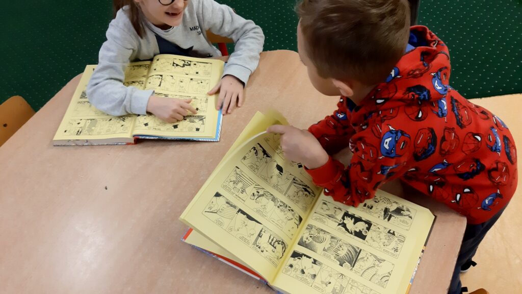 Zdjęcie przedstawia dwoje dzieci chłopca i dziewczynkę siedzących przy stoliku. Przed nimi leżą otwarte komiksy.