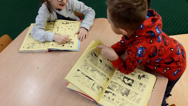 Zdjęcie przedstawia dwoje dzieci chłopca i dziewczynkę siedzących przy stoliku. Przed nimi leżą otwarte komiksy.