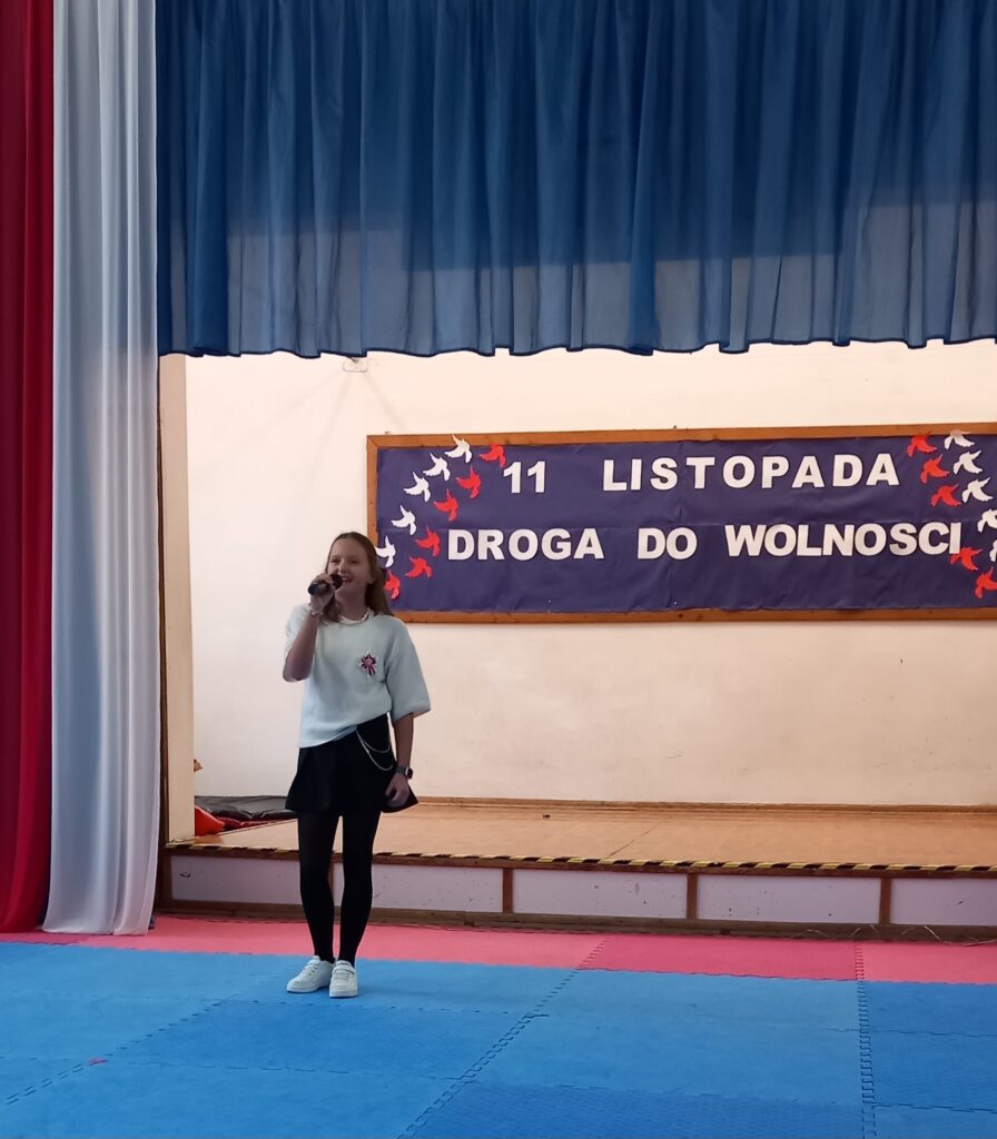 na rozświetlonej scenie uczestniczka ubrana w strój galowy trzyma mikrofon. W tle tablica okolicznościowa z napisem :11 listopada - droga do wolności"