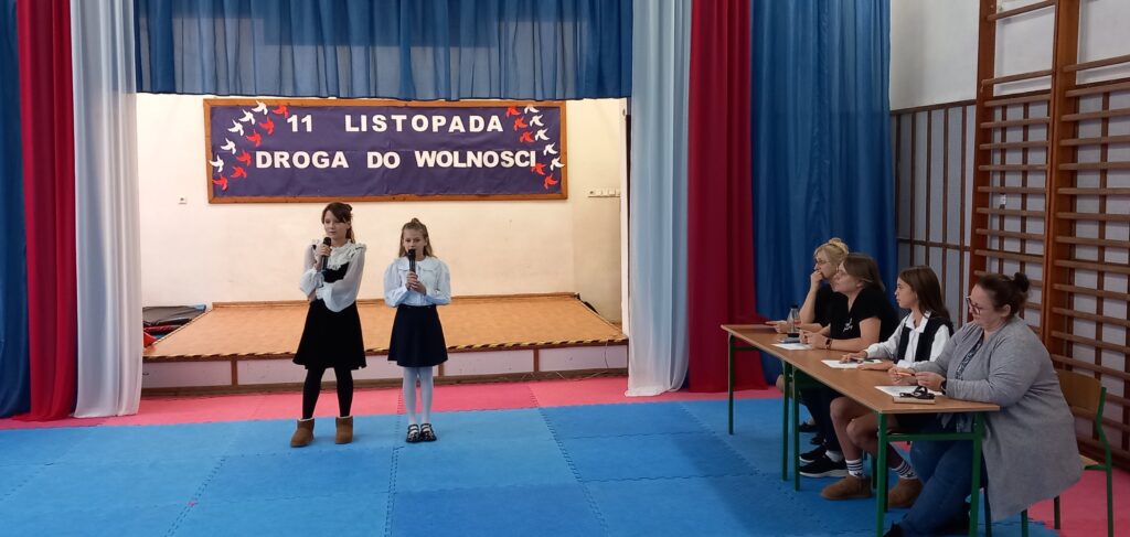 na rozświetlonej scenie okolicznościowa z napisem :11 listopada - droga do wolności"dwie uczestniczki z klasy 4, ubrane w strój galowy, trzymają mikrofony. Po prawe stronie jury - 3 osoby i prowadząca konkurs