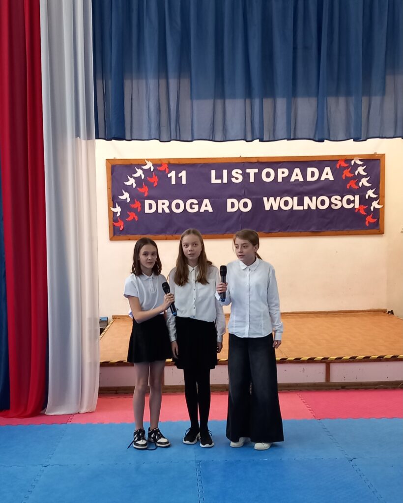 na rozświetlonej scenie trzy uczestniczki ubrane na galowo stoją na środku sceny, skrajne trzymają mikrofony.