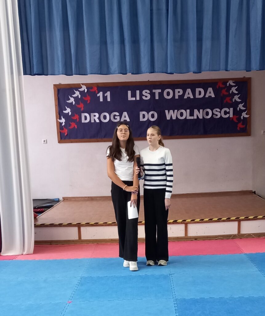 na środku oświetlonej sceny stoją dwie dziewczynki, ubrane w strój galowy, jedna trzyma mikrofon