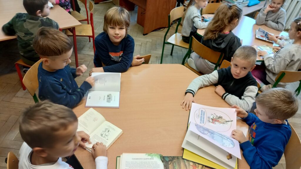 zdjęcie pokazuje grupę dzieci siedzących przy stoliku w bibliotece i przeglądających książki. Dzieci są zainteresowane swoimi lekturami, a na stole leży kilka otwartych książek, z których jedna zawiera ilustracje.