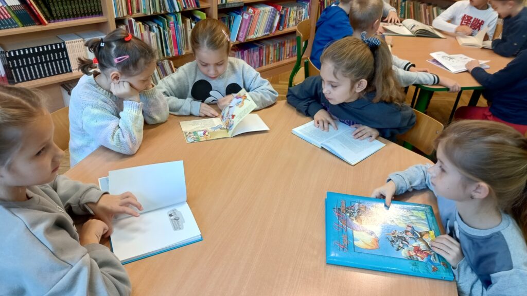 zdjęcie przedstawia dzieci siedzące przy stoliku w bibliotece, przeglądające różne książki. Każde dziecko skupia się na swojej książce, a niektóre z nich mają kolorowe ilustracje. Wystrój otoczenia pełen jest półek z książkami, co podkreśla atmosferę biblioteki.