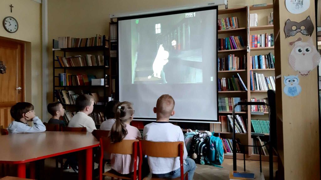 Zdjęcie przedstawia biały ekran rozciągnięty na tle półek z książkami. Na ekranie widać wyświetlaną animację. Przed ekranem siedzą na krzesełkach dzieci. Za nimi stoi stół.