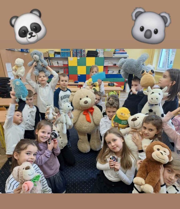 Na zdjęciu widzimy grupę dzieci w szkolnej klasie, które pozują z różnymi pluszowymi maskotkami. W centralnej części stoi duży pluszowy miś z pomarańczową kokardą. Dzieci są uśmiechnięte i trzymają w rękach pluszowe zwierzątka, takie jak panda, małpa, owca czy krowa. W tle widać półki z książkami oraz kolorowe szafki, typowe dla sali lekcyjnej. Atmosfera jest radosna, co sugeruje, że jest to specjalne wydarzenie, być może Dzień Pluszowego Misia.