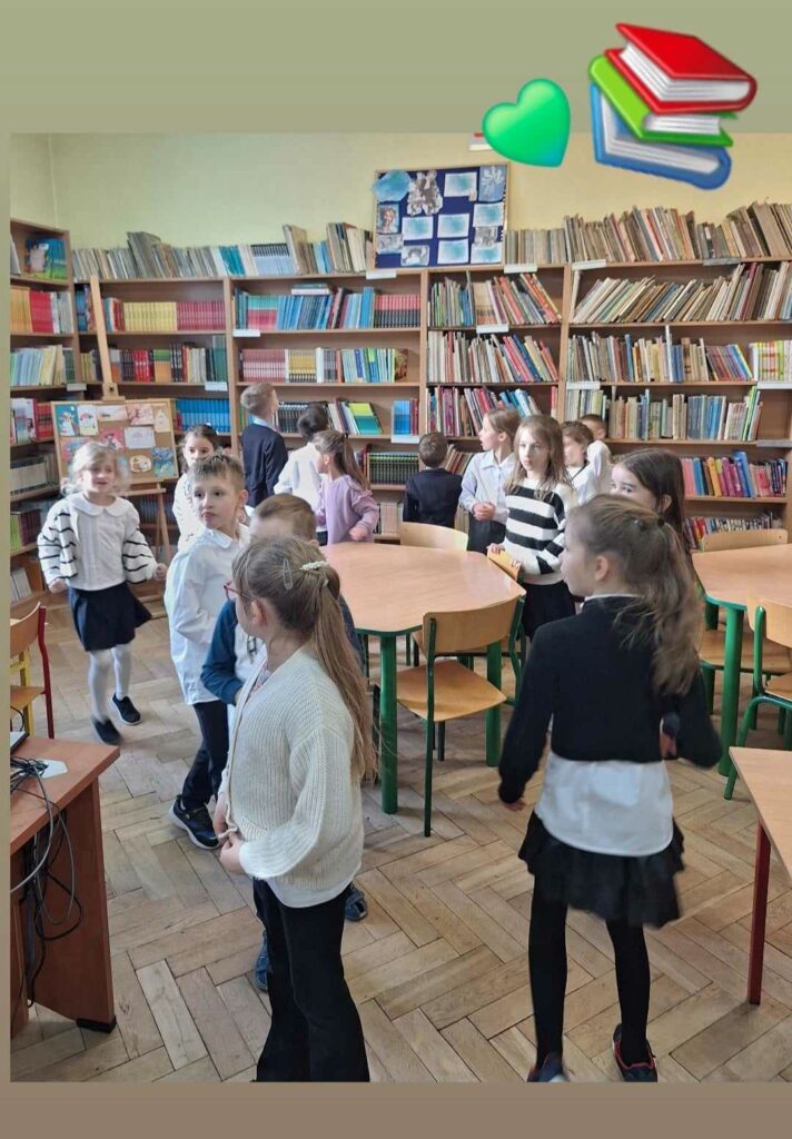 Na zdjęciu widać grupę dzieci w szkolnej bibliotece. Otaczają ich regały pełne książek o różnokolorowych okładkach. Dzieci ubrane są w schludne stroje, a atmosfera wydaje się radosna i dynamiczna. Na pierwszym planie widoczne są stoły i krzesła, które tworzą przestrzeń do nauki lub zabawy. W tle można dostrzec tablicę z pracami plastycznymi, która dodaje wnętrzu charakteru.