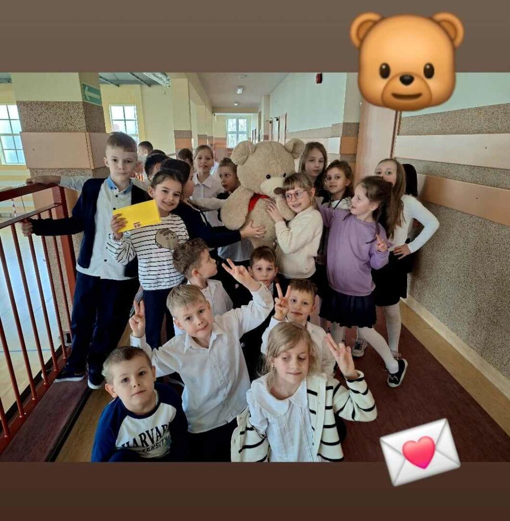 Zdjęcie przedstawia grupę dzieci stojących w korytarzu szkoły, uśmiechających się i pozujących do zdjęcia. W centrum uwagi znajduje się duży pluszowy miś, którego kilka dzieci przytula. Dzieci są ubrane w eleganckie stroje, co może wskazywać na uroczystą okazję lub specjalny dzień w szkole. Na górze zdjęcia dodano emotikony: misia oraz kopertę z sercem, co podkreśla radosny i przyjazny charakter sceny. Korytarz szkolny jest jasny, z dużymi oknami, a w tle widać inne dzieci oraz elementy szkolnego wyposażenia.