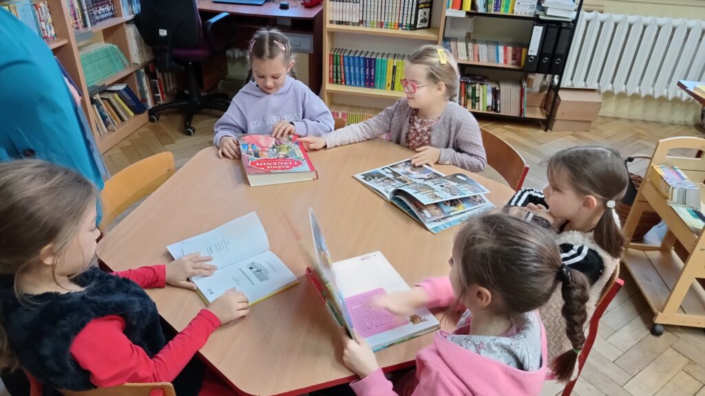 Zdjęcie przedstawia dziewczynki siedzące przy sześciokątnym stoliku. Dziewczynki oglądają różne książki. W tle widać regały z książkami.